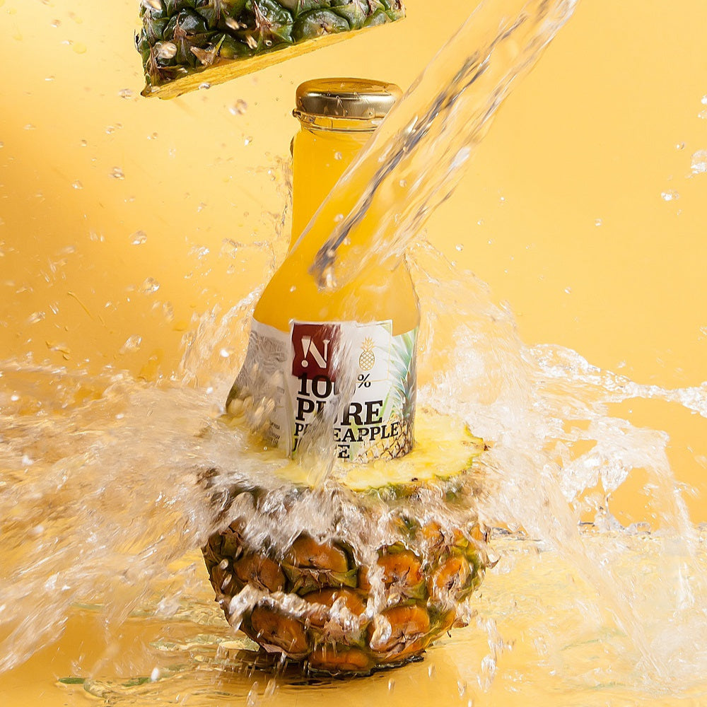 Bottle of pineapple juice being poured into a pineapple with water splashing around on a yellow background